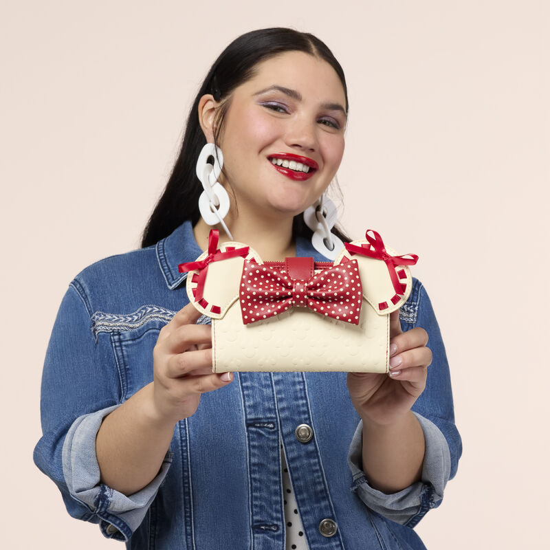 Woman smiling at camera wearing a white dress with black polka dots and denim jacket. She holds the Loungefly Minie Mouse Rock the Dots wallet in both hands and holds it up to camera. 